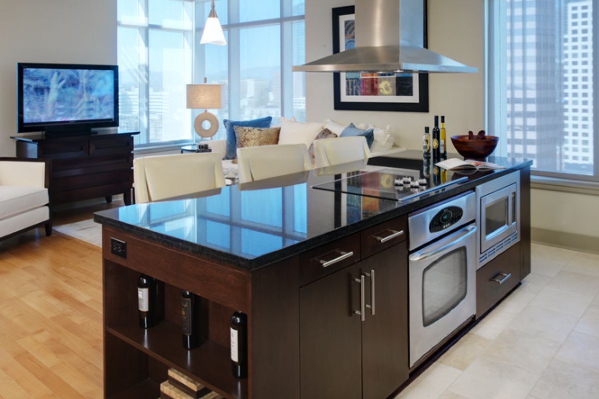 Modern kitchen with a glossy island, built-in oven, and a view of a bright, open living area with large windows.