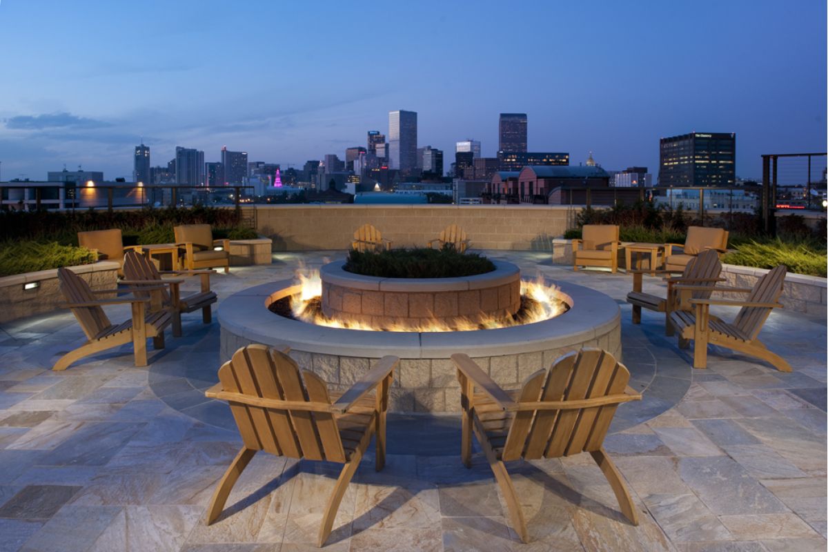 Wooden chairs surround a circular firepit on a rooftop patio with a city skyline in the background at dusk.