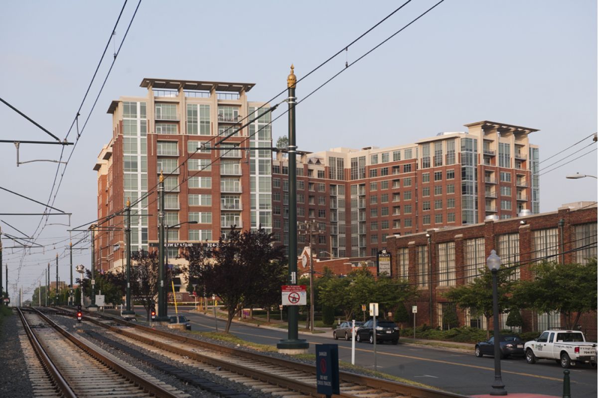 Apartment buildings and trees beside train tracks and street, with overhead power lines visible.