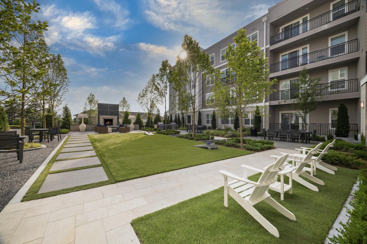 Modern apartment courtyard with green lawn, white chairs, trees, paved pathways, and a fireplace under a blue sky.