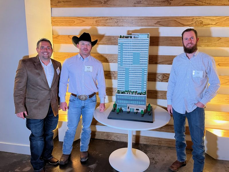 Three men stand by a detailed scale model of a tall building on a round table, against a wood-panel wall.