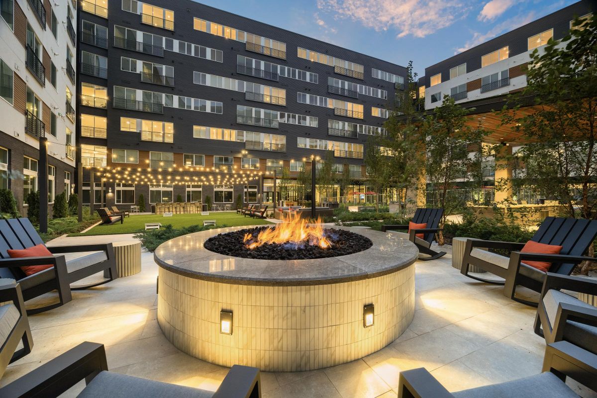 Modern courtyard with a large circular fire pit, surrounded by chairs, string lights, and a multi-story building.