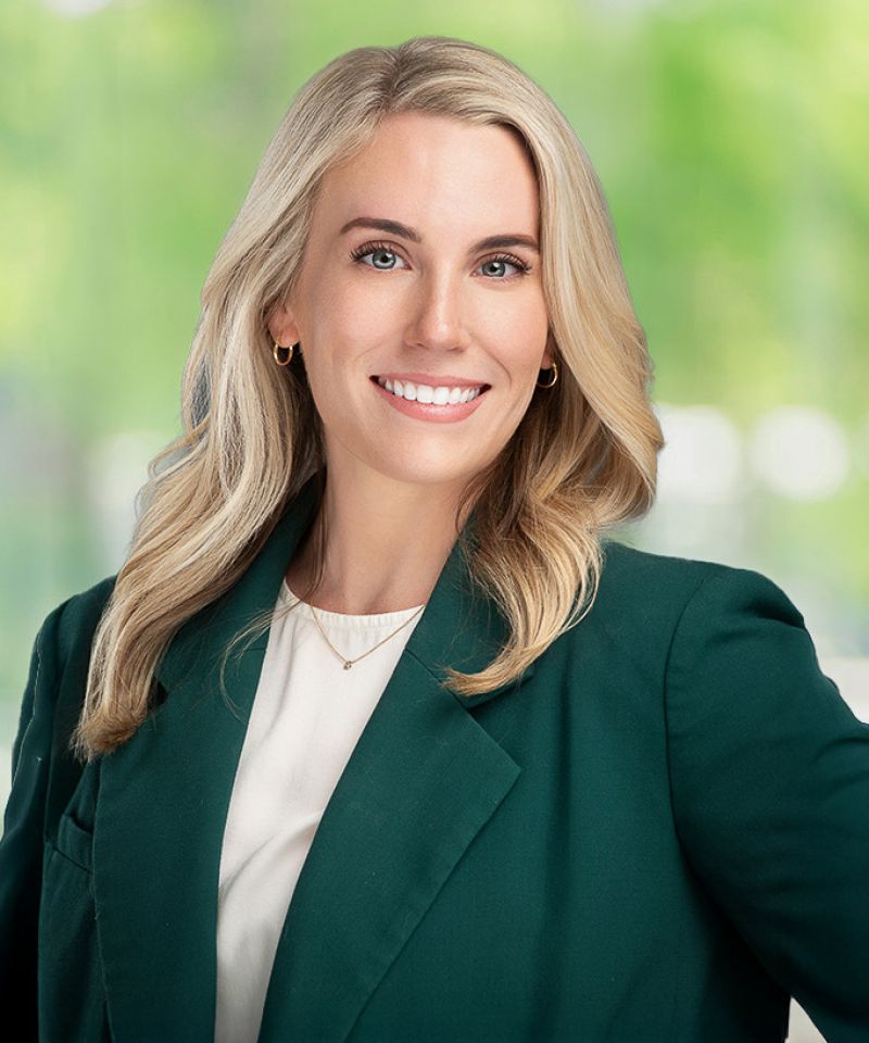 Caroline Erck, a smiling woman with long blonde hair in a green blazer and white top, stands before a blurred background.