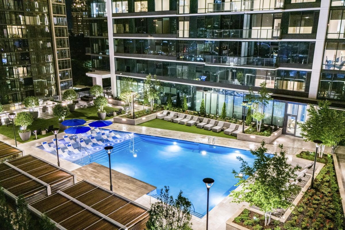 Night view of a modern apartment building with a lit outdoor swimming pool, surrounded by greenery and lounge chairs.