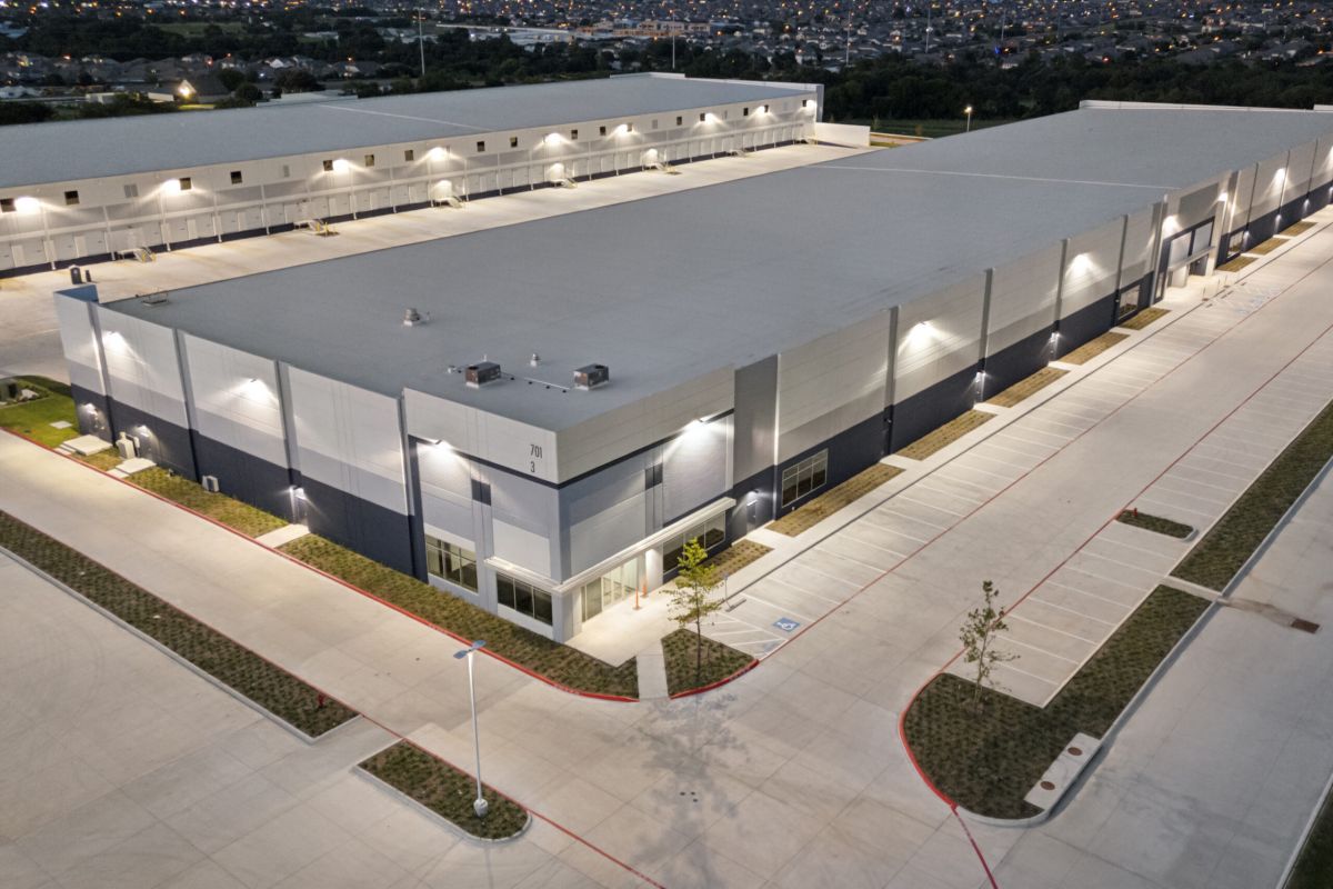 A large, modern warehouse building with exterior lights on and an empty parking lot at dusk.