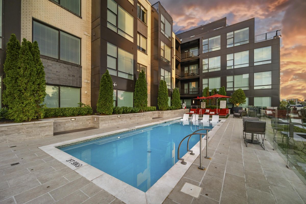 Modern apartment building with a pool, surrounded by trees and lounge chairs, under a vibrant sunset sky.
