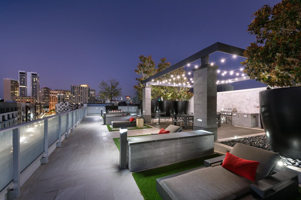 Modern rooftop lounge at dusk with city views, string lights, lounge chairs, and red-accented pillows.