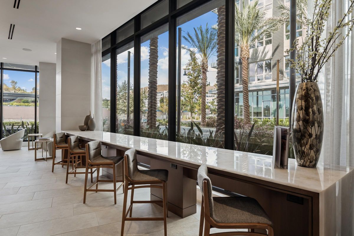 Hanover Laguna Niguel Modern lounge area with a long table, chairs, and large windows overlooking a courtyard with palm trees.