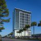 Tall modern building with glass facade, surrounded by palm trees and a clear blue sky. Street view with parked cars.