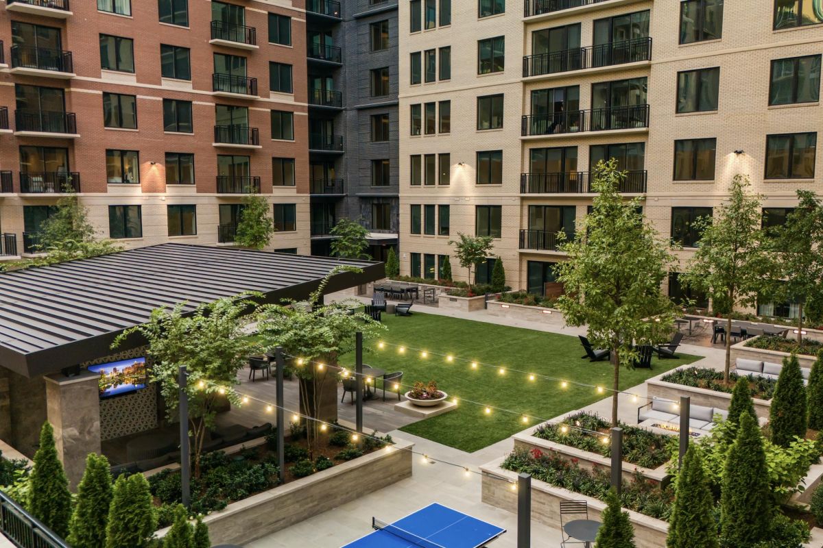 Apartment courtyard with a lawn, seating, tables, string lights, and a ping pong table surrounded by trees and buildings.