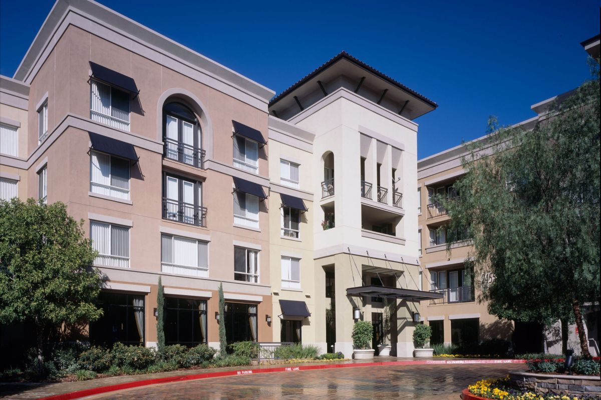 Four-story modern hotel with balconies, large windows, and landscaping under a clear blue sky.