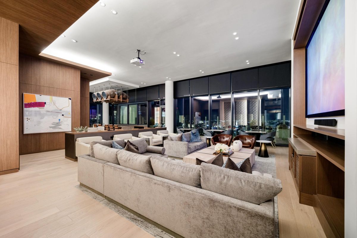 Modern living room with plush sectional sofa, abstract art, projector screen, and large windows showing city view.