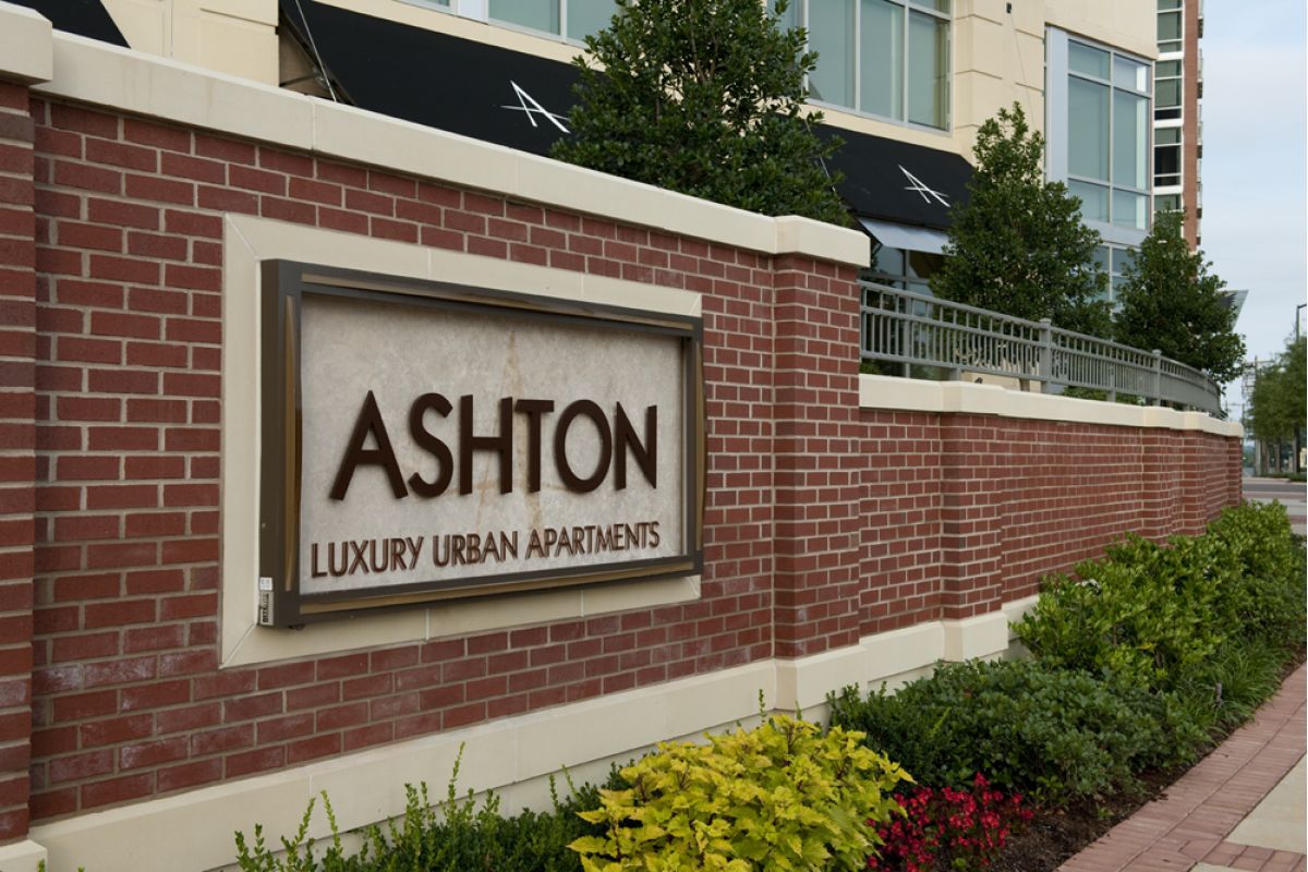 A brick sign reads "ASHTON LUXURY URBAN APARTMENTS" in front of a modern apartment building and plants.
