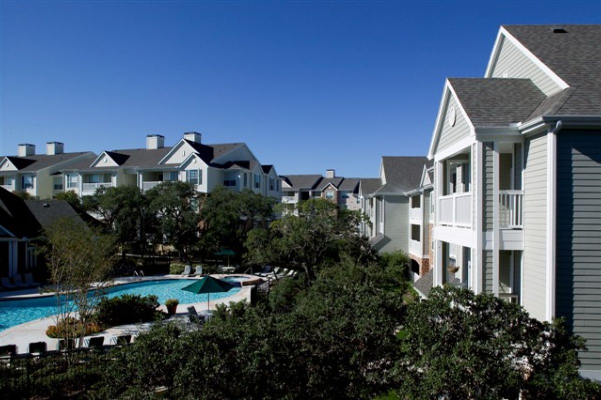 Apartment buildings with balconies overlook a swimming pool and trees on a clear, sunny day.