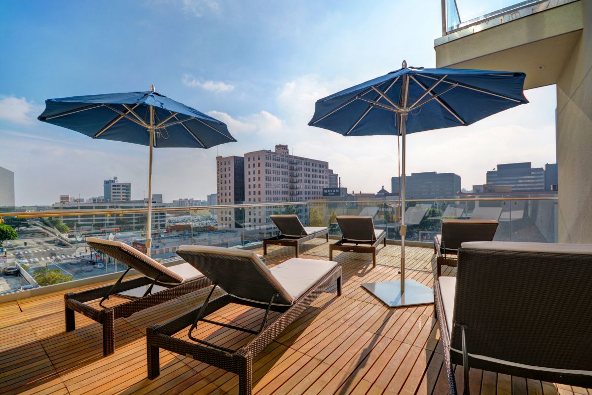 Rooftop deck with lounge chairs and blue umbrellas overlooking a city skyline on a sunny day.