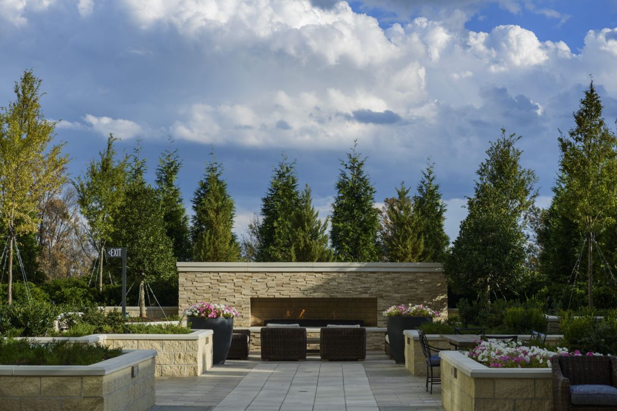 Outdoor patio with wicker chairs, flower planters, stone fireplace, and trees under a partly cloudy sky.