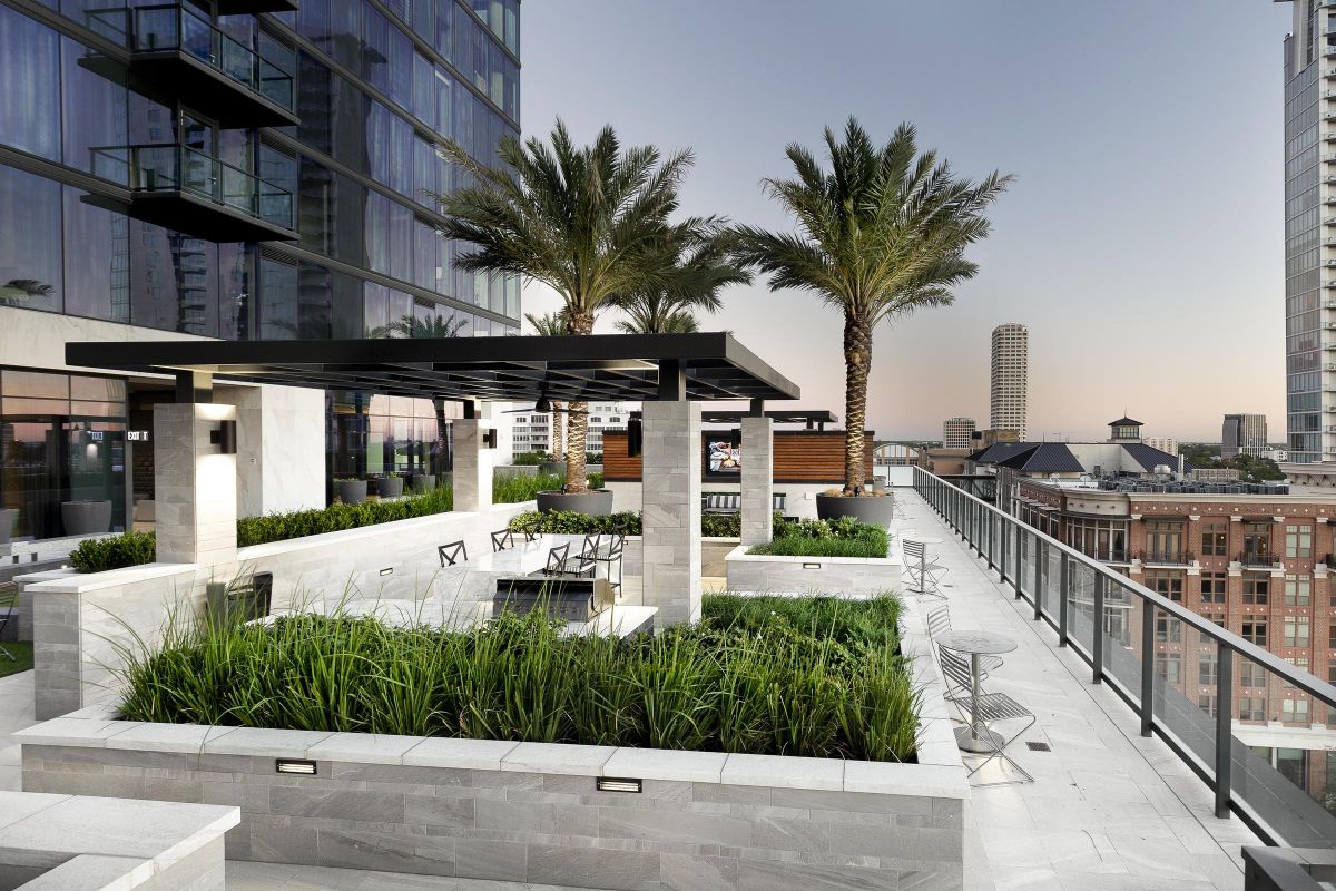 Modern rooftop terrace with palm trees, greenery, and seating areas overlooking a cityscape at dusk.