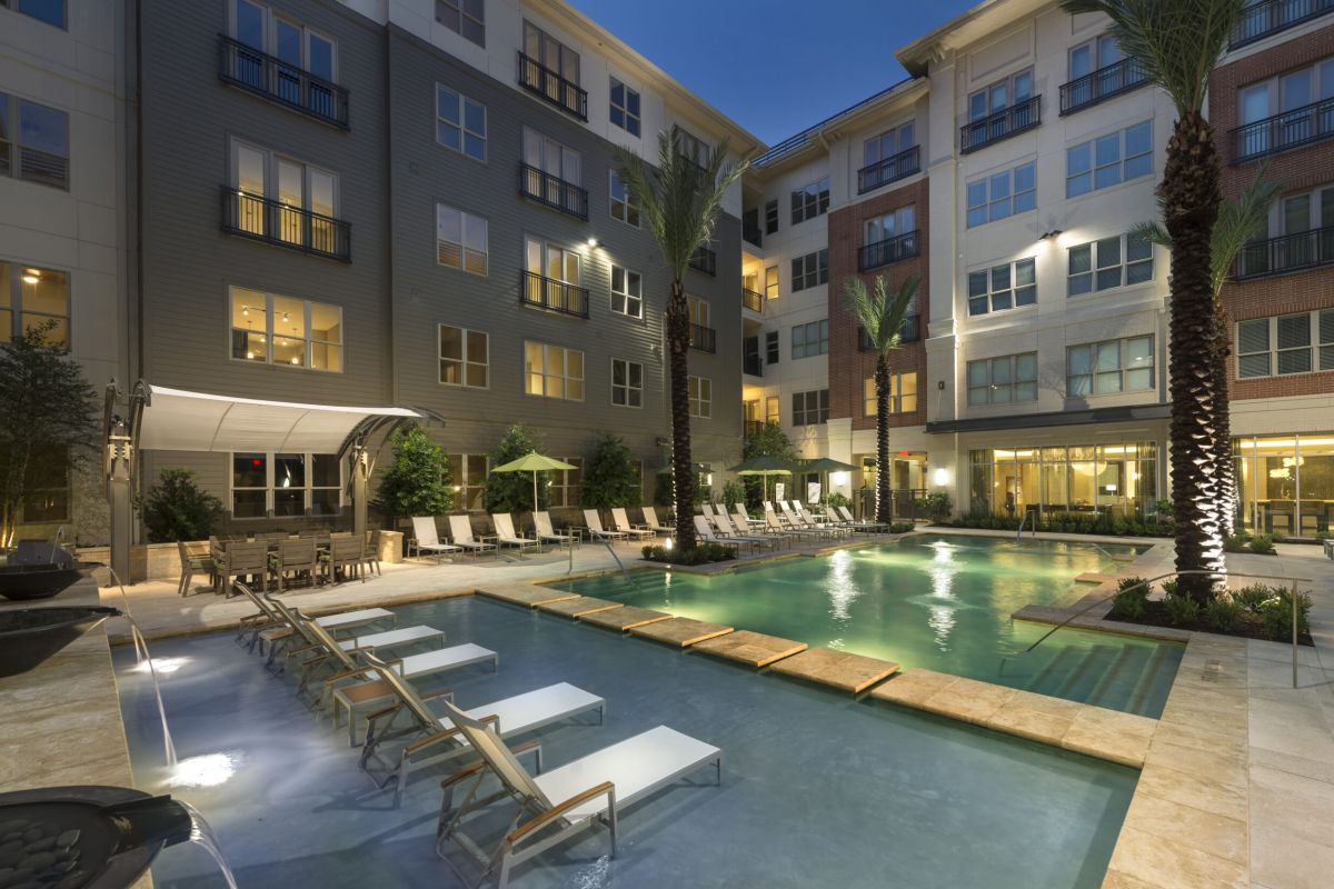 Modern apartment complex courtyard with a lit swimming pool, lounge chairs, and tall palm trees at dusk.