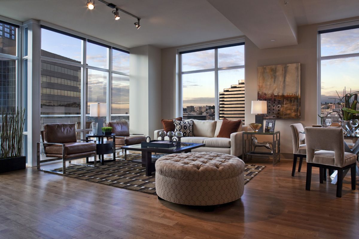 Modern living room with large windows, city view, leather chairs, round ottoman, and dining area.