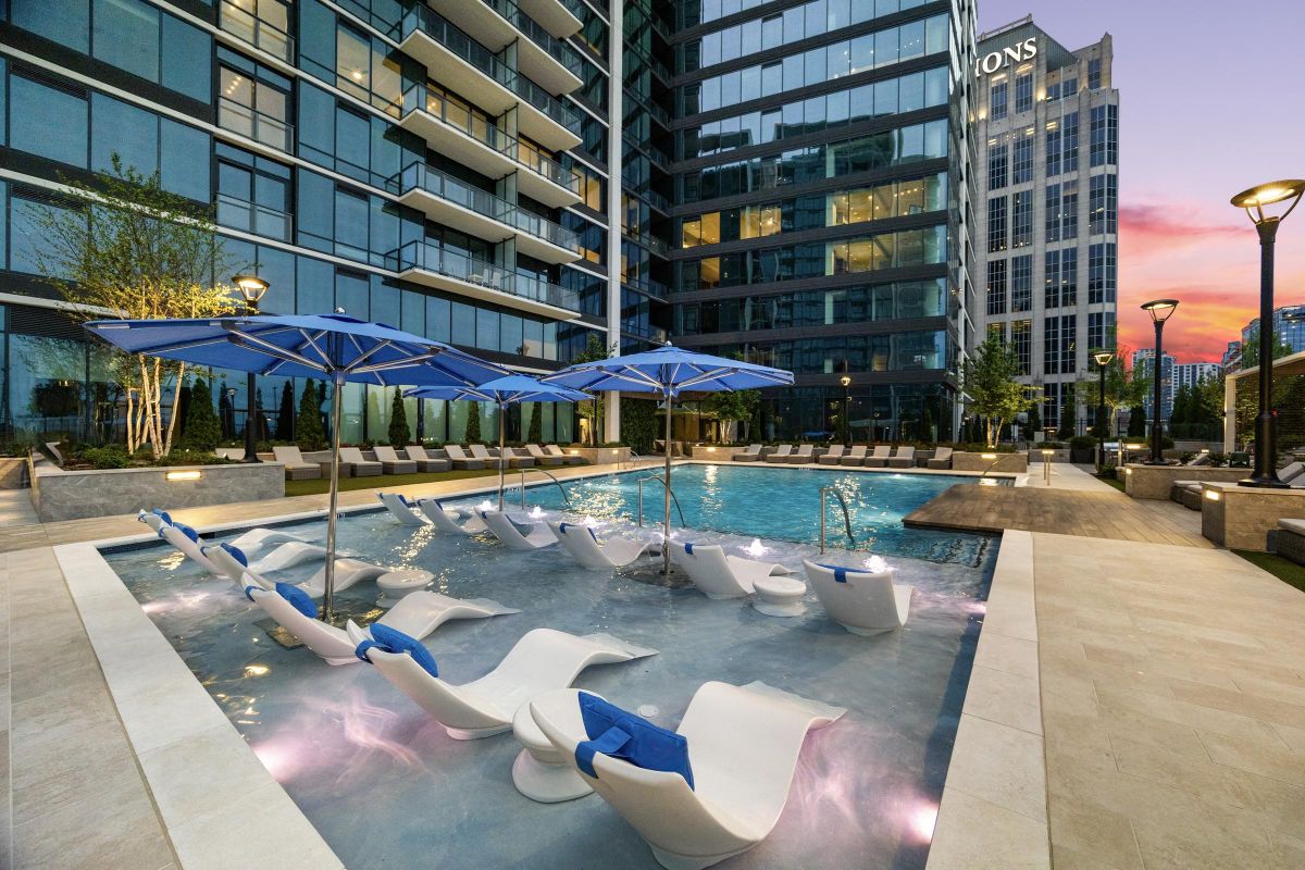 Modern pool area with lounge chairs in shallow water, blue umbrellas, and surrounding high-rise buildings at sunset.