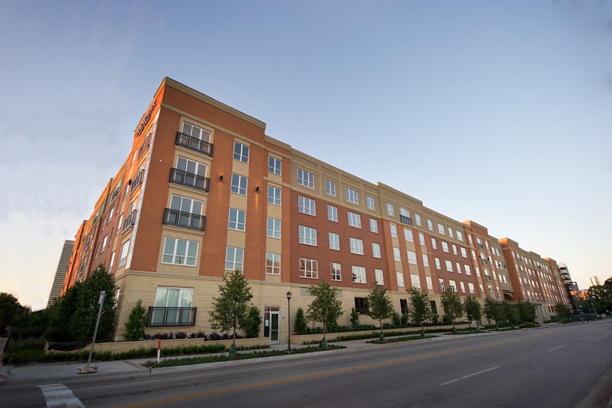 A modern, multi-story apartment building beside a quiet street lined with trees under a clear sky.
