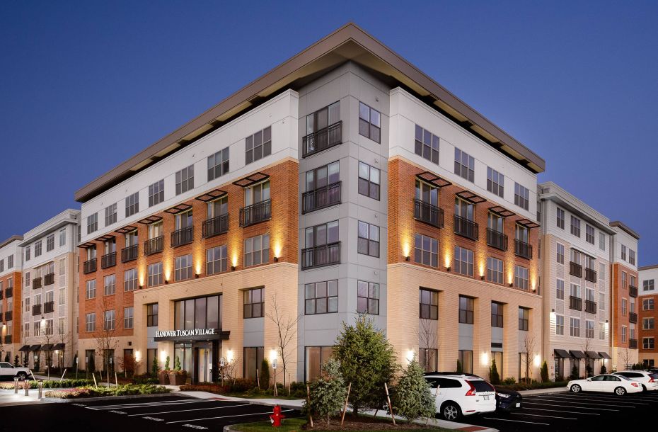 Hanover Company, Hanover Tuscan Village Modern apartment building with illuminated facade at dusk, surrounded by parked cars and landscaping.