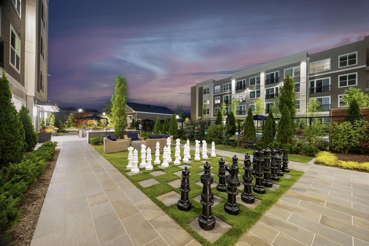 Rooftop garden with giant chess set, surrounded by modern buildings, under a twilight sky.