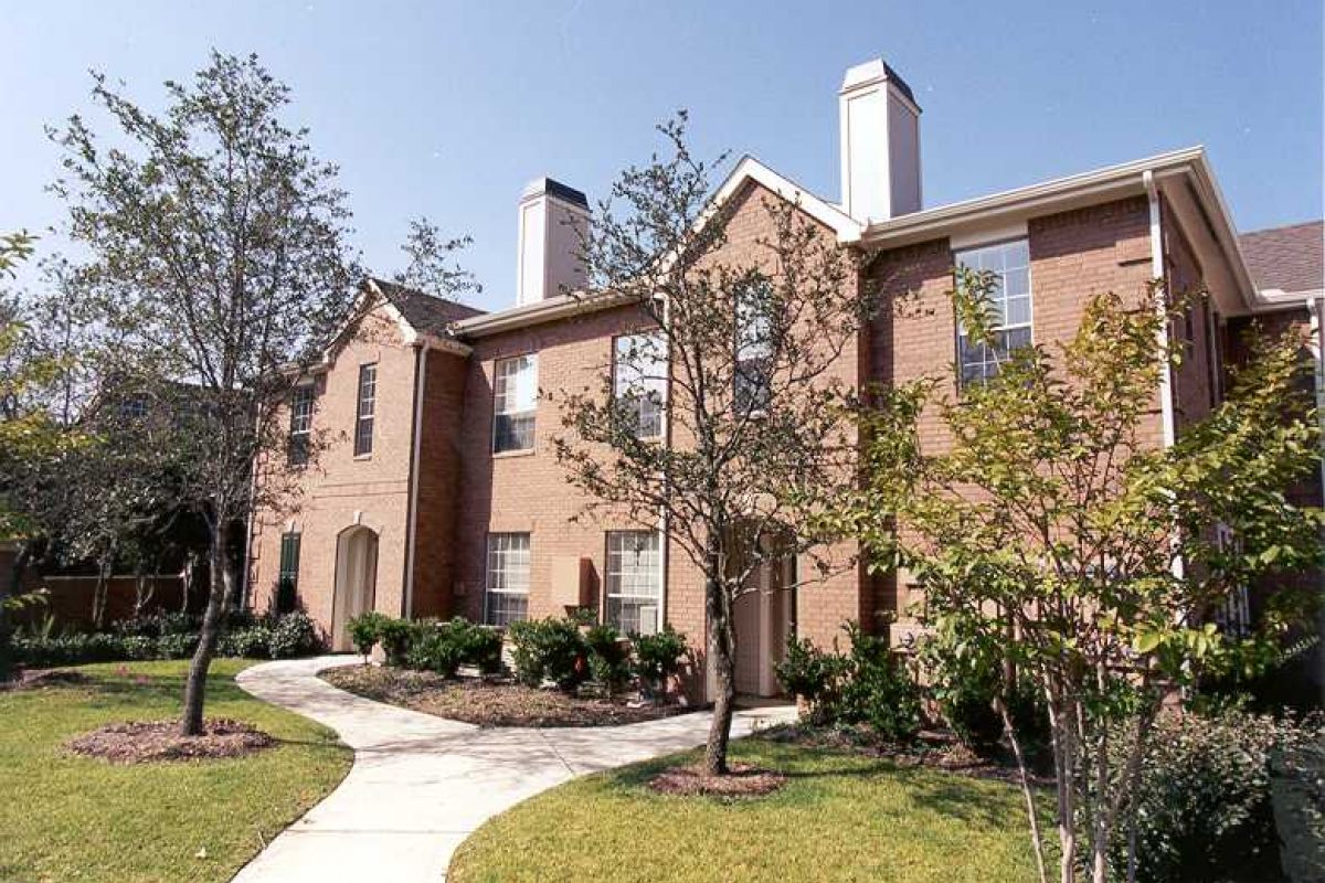 Two-story brick Churchill on the Park apartments with chimneys, trees, and a curved sidewalk in sunny Dallas.