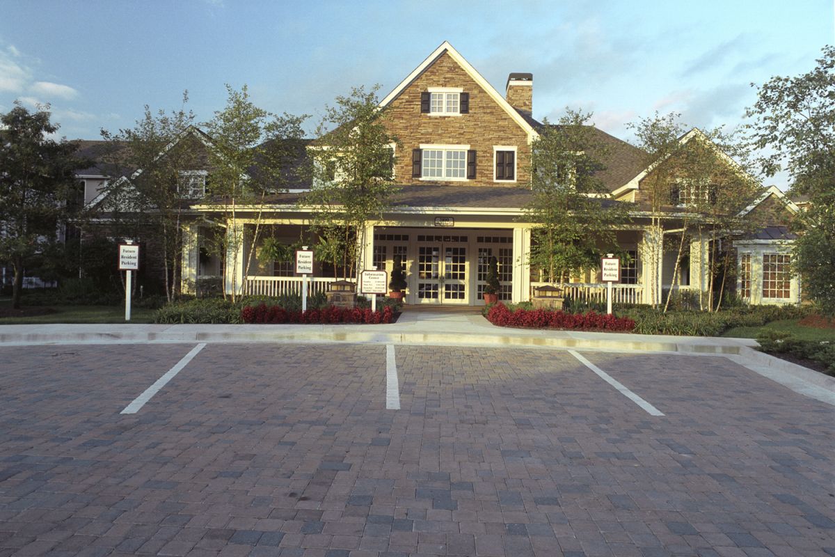 A brick building with white trim, porch, and parking spaces in front, surrounded by trees and flowers.