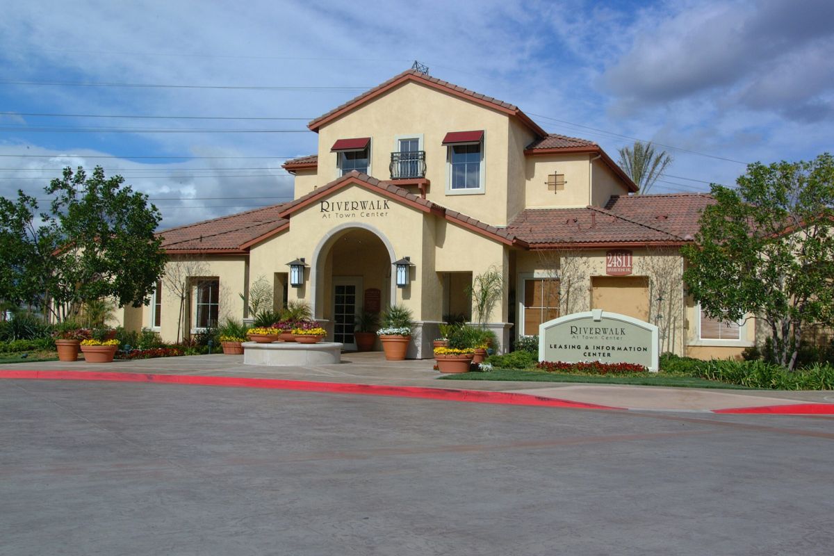 A tan building with red trim reading “Riverwalk” and surrounded by plants under a cloudy sky.