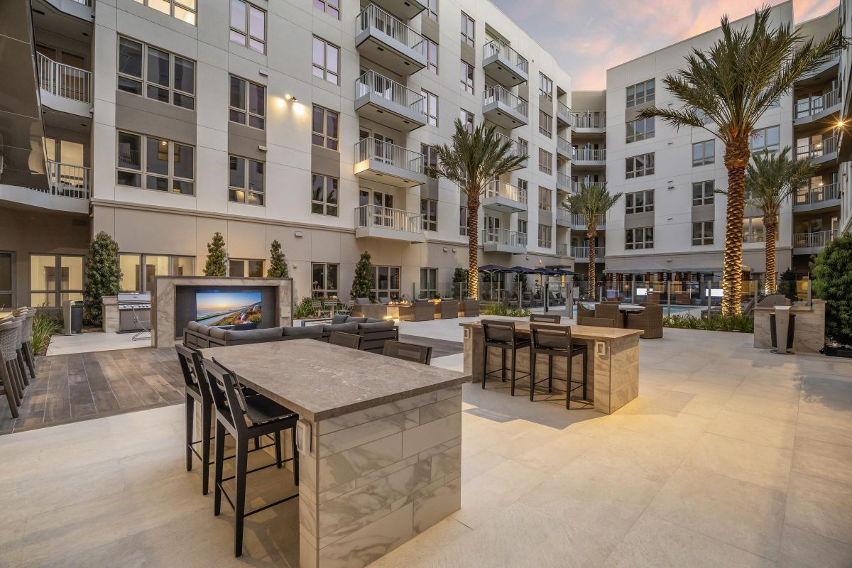 Modern apartment courtyard with outdoor seating, tables, and palm trees, set under a colorful evening sky.