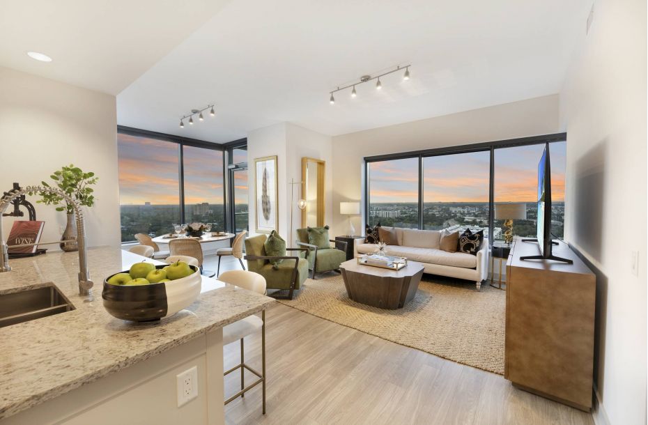 Hanover Company, The Driscoll at River Oaks Modern open-plan living room with large windows, cozy seating, a kitchen island, and city sunset views.