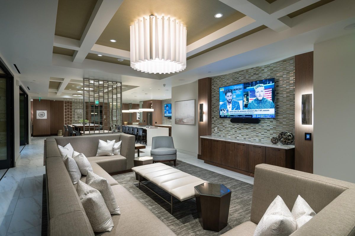 Modern lounge with gray sofas, a TV on the wall, a large chandelier, and a sleek kitchen in the background.