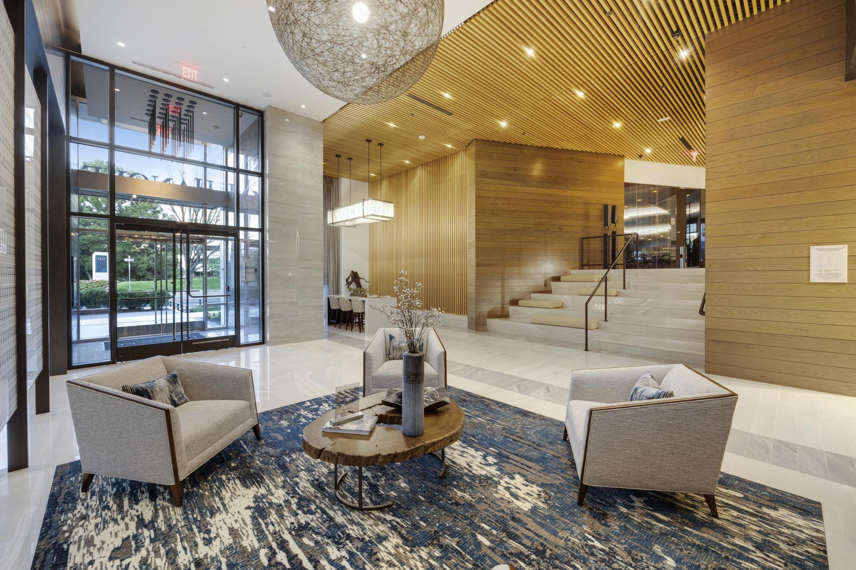 Modern lobby with two grey chairs, wooden table, blue rug, and large windows.