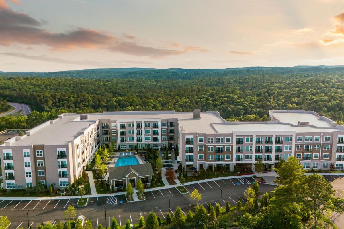 A modern apartment complex with a central pool, surrounded by trees and mountains under a cloudy sky.