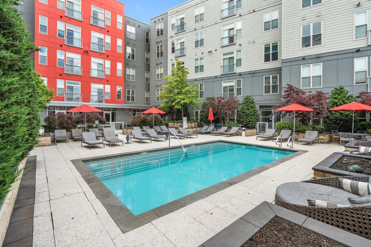 Apartment complex pool area with lounge chairs, red umbrellas, and surrounding plants.