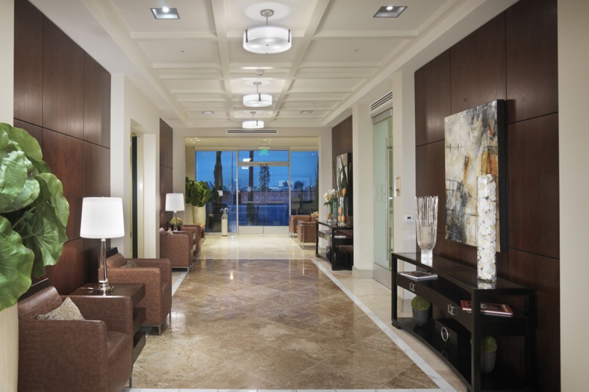 Modern office lobby with brown chairs, plants, artwork, and large windows showing a cityscape at dusk.