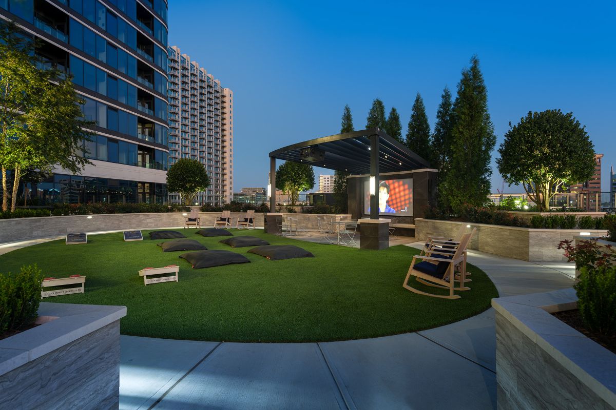 Modern outdoor lounge area with chairs, grass, and a covered screen, surrounded by tall buildings at dusk.
