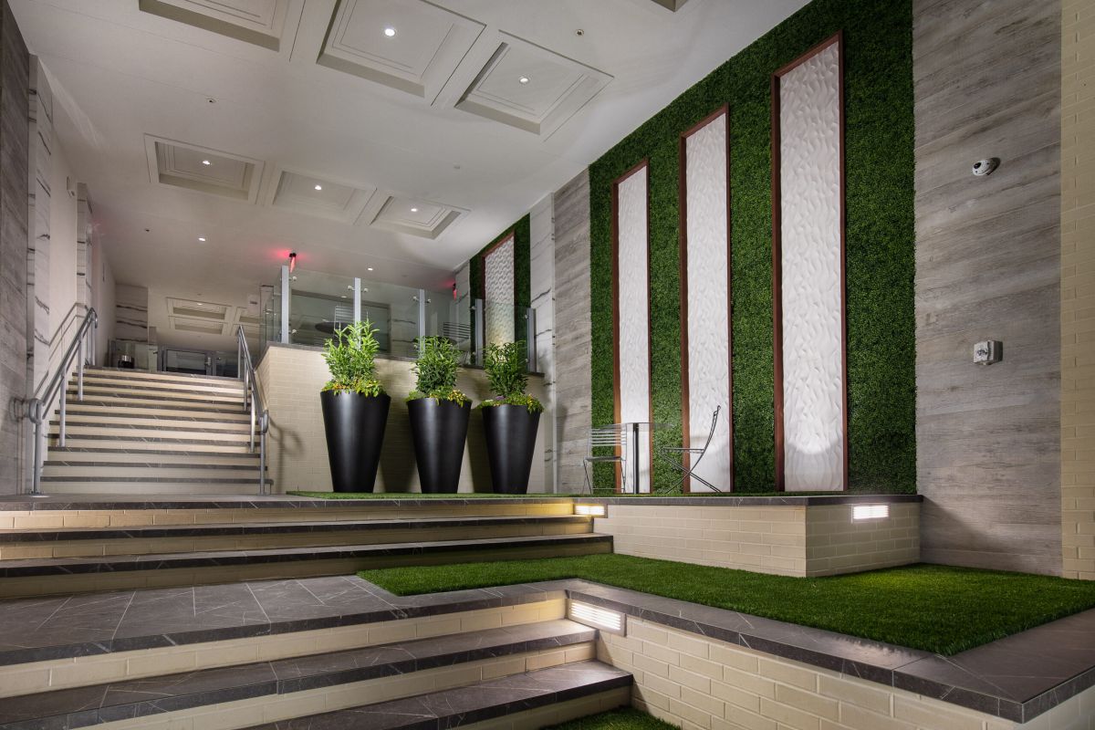 Modern indoor lobby with green wall panels, tall potted plants, and tiered steps with artificial grass.