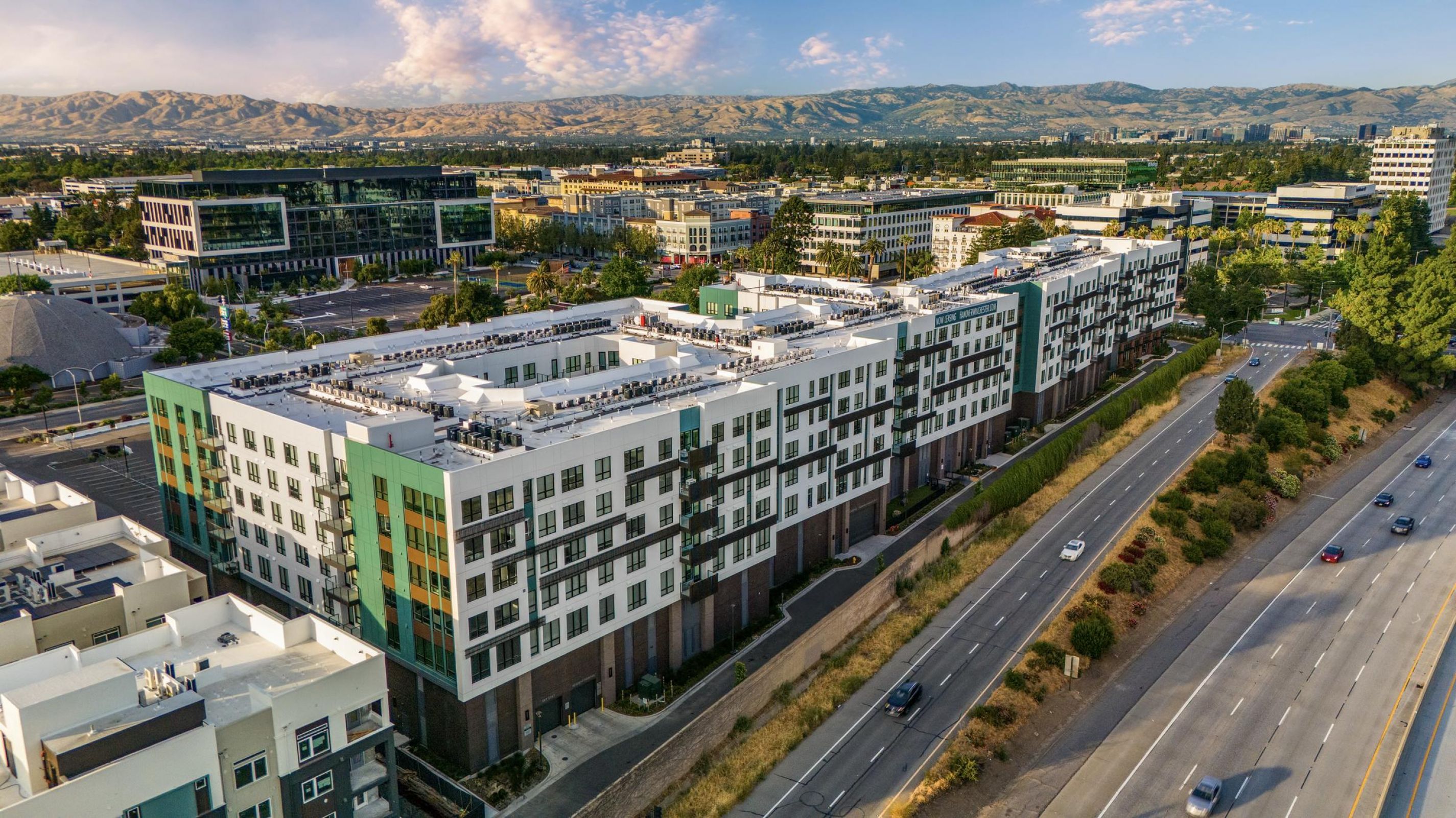 Aerial view of urban buildings with a highway nearby, mountains, and a cloudy sky in the background.