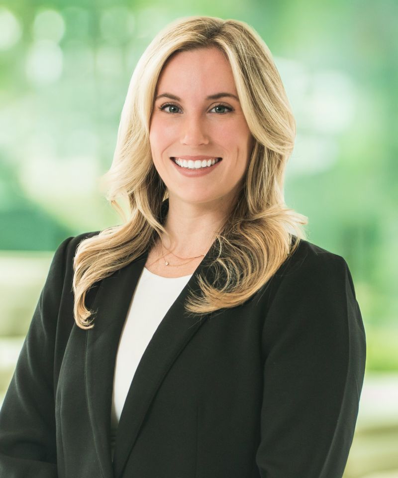 Smiling woman with long blonde hair in a black blazer, standing in front of a blurred green background.