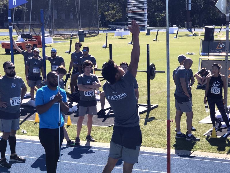 Man jumps to touch a vertical measuring pole during an outdoor athletic event; others watch and cheer.