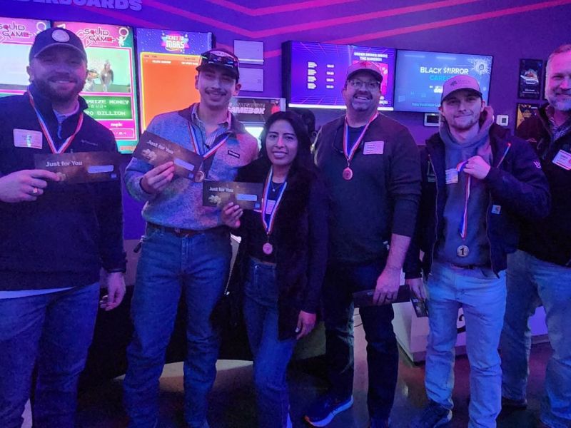 Six people wearing medals pose indoors, holding cards and smiling in a brightly lit gaming venue.