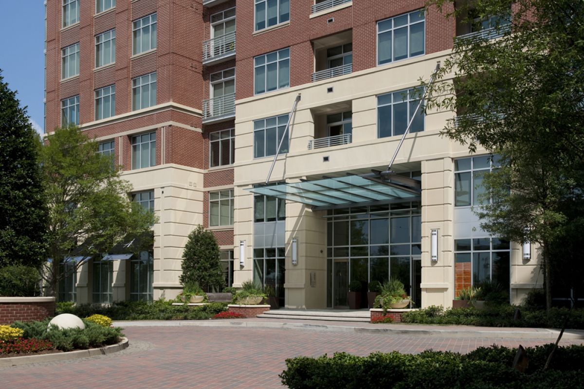 Modern apartment building entrance with glass canopy, potted plants, and landscaped brick driveway.