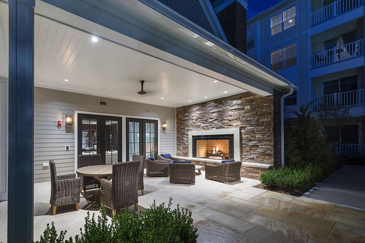 Covered patio at night with wicker chairs, round table, and a stone fireplace lit with a cozy fire.