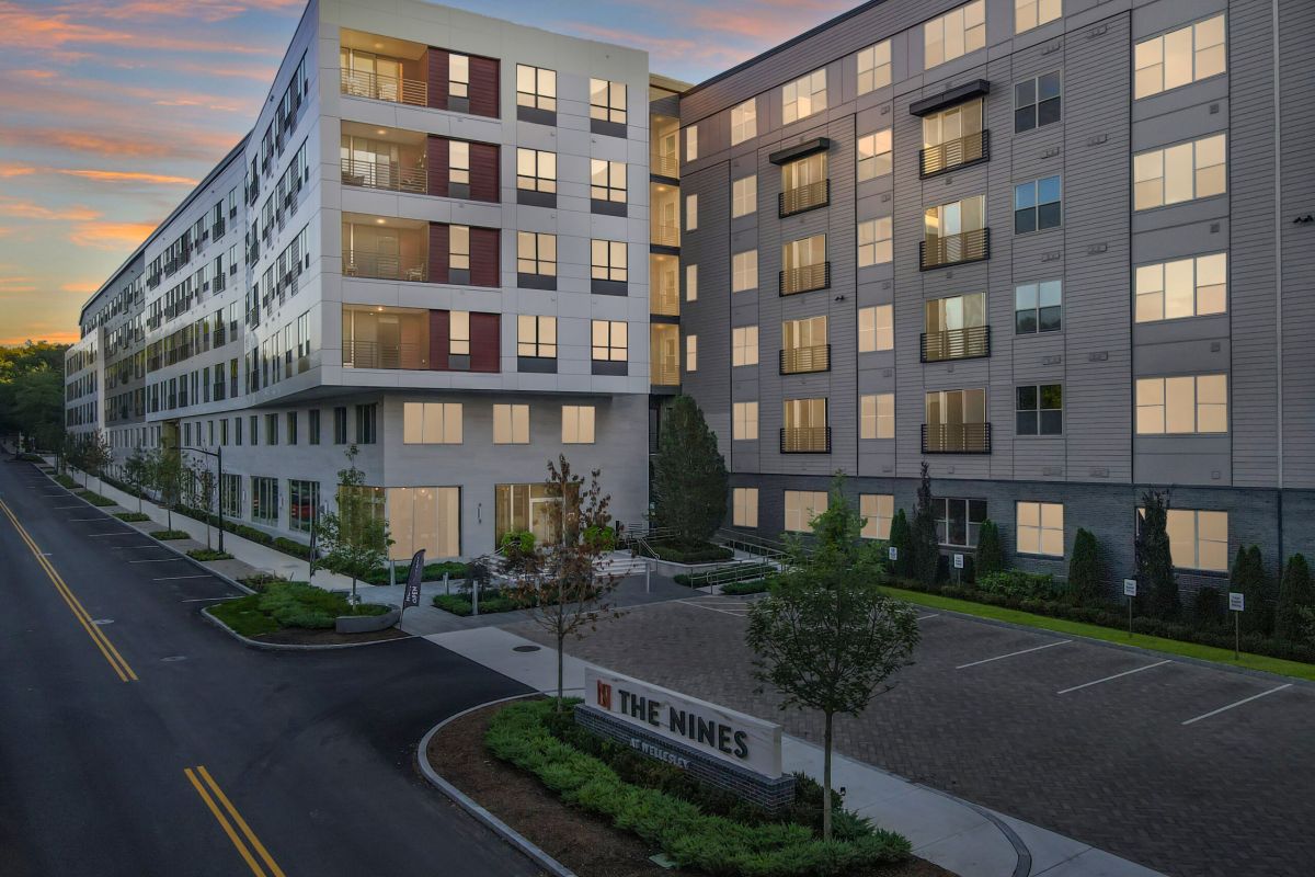 Modern apartment building called "The Nines" at sunset, with lit windows and empty street in front.
