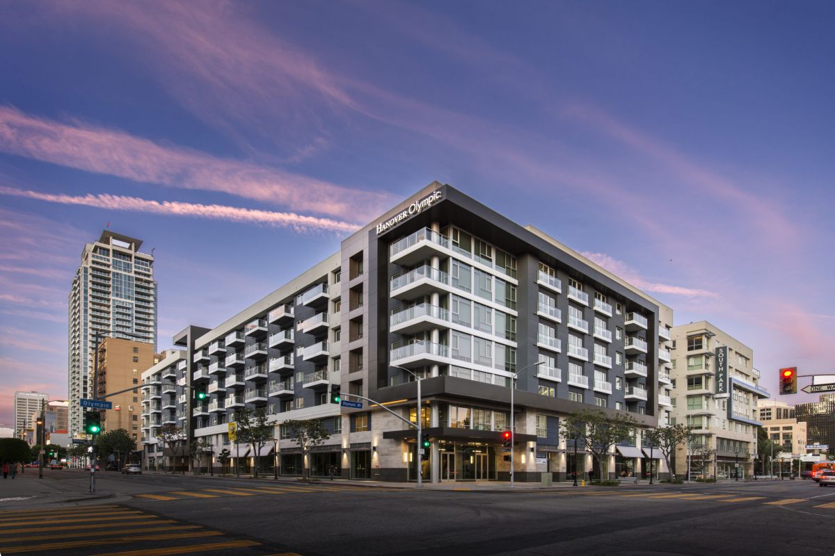 Modern apartment building on a city corner at sunset, with pink clouds and a tall building in the background.