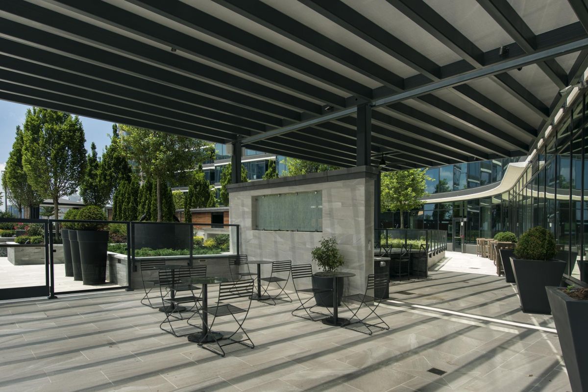 Modern outdoor patio with metal tables and chairs, potted plants, and glass partitions under a slatted pergola.