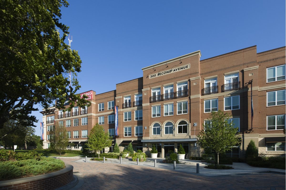 Four-story red brick apartment at Charles River Landing features balconies, arched entries, and trees out front on a sunny day.