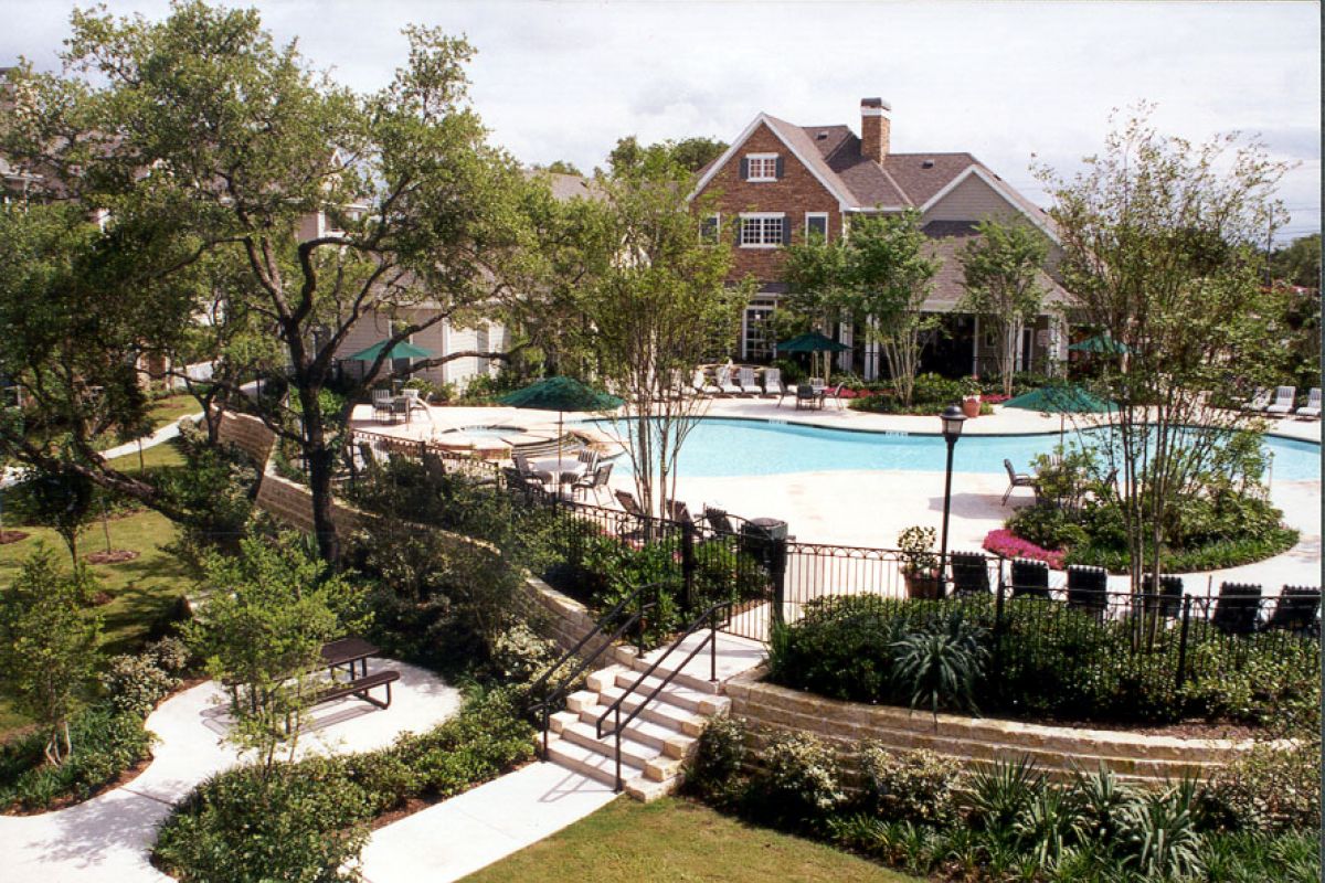 A landscaped outdoor pool area with lounge chairs, trees, and a large house in the background.